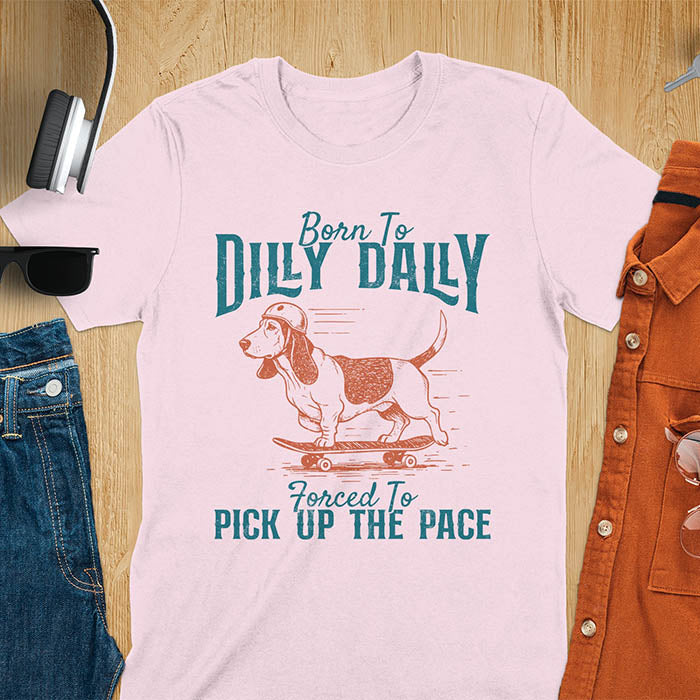 Basset hound wearing helmet on skateboard with Born To Dilly Dally Forced To Pick Up The Pace lettering on light pink tee