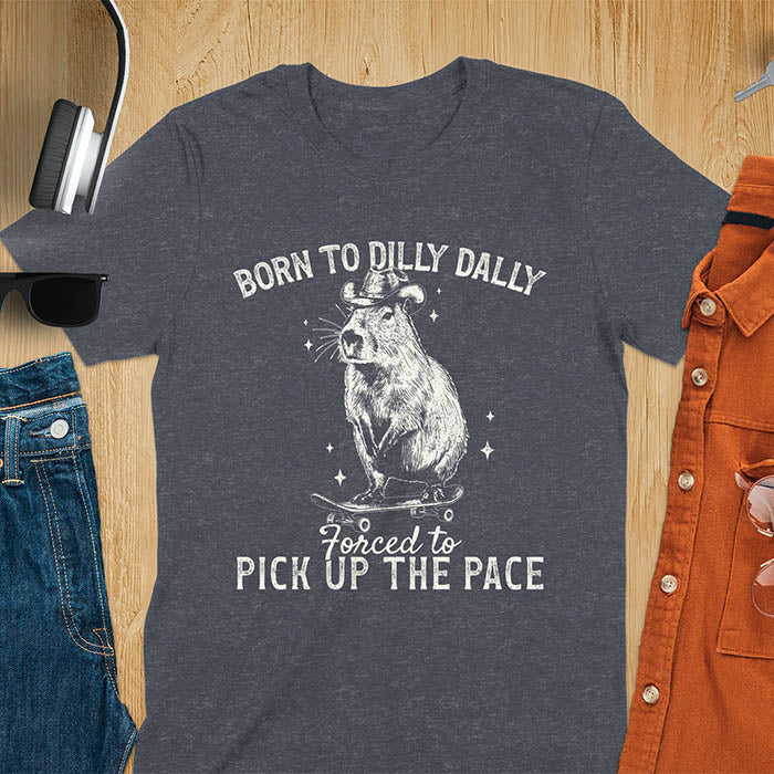 Dark heather color tee featuring aCapybara in a cowboy hat on a skateboard. Born to Dilly Dally and Forced to Pick Up the Pace text. Distressed style.