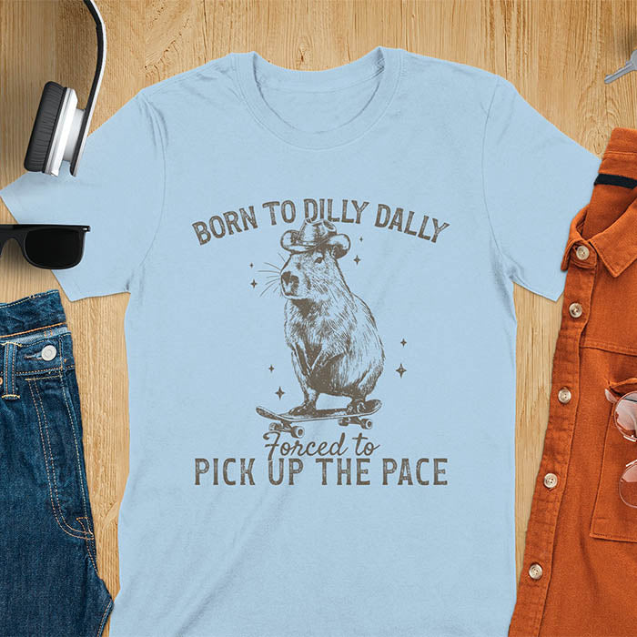 Light blue color tee featuring a Capybara in a cowboy hat on a skateboard. Born to Dilly Dally and Forced to Pick Up the Pace text. Distressed style.