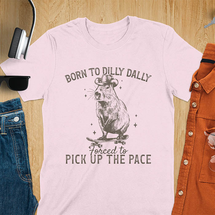 Light pink color tee featuring a Capybara in a cowboy hat on a skateboard. Born to Dilly Dally and Forced to Pick Up the Pace text. Distressed style.