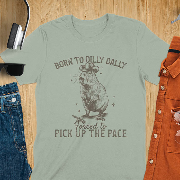 Sage color tee featuring aCapybara in a cowboy hat on a skateboard. Born to Dilly Dally and Forced to Pick Up the Pace text. Distressed style.