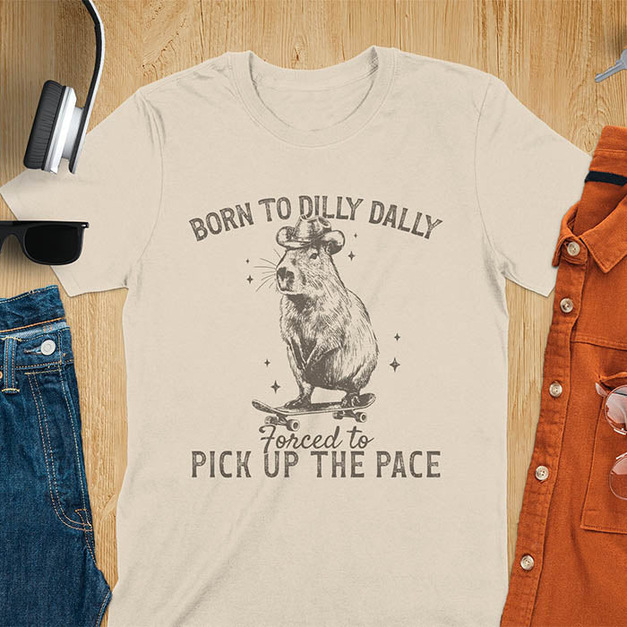 Sand color tee featuring a Capybara in a cowboy hat on a skateboard. Born to Dilly Dally and Forced to Pick Up the Pace text. Distressed style.