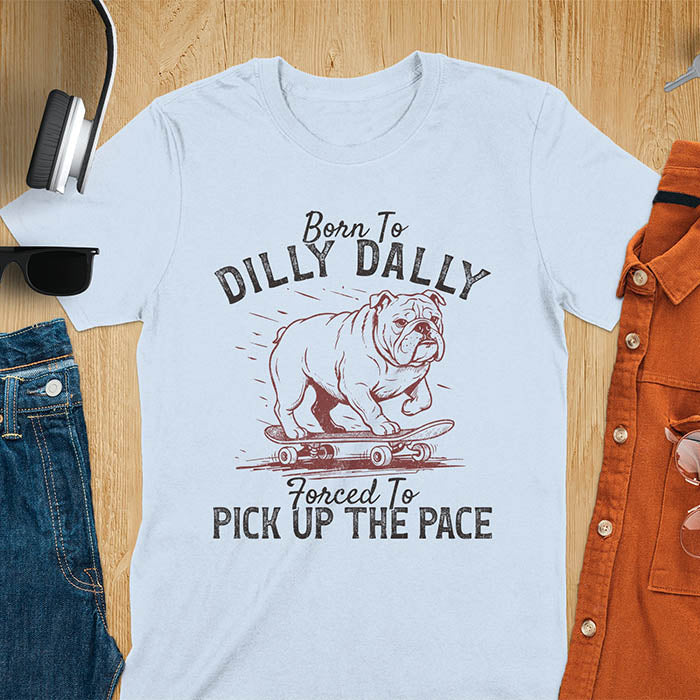Grumpy bulldog on skateboard illustration with Born To Dilly Dally text and Forced To Pick Up The Pace slogan on light blue tee
