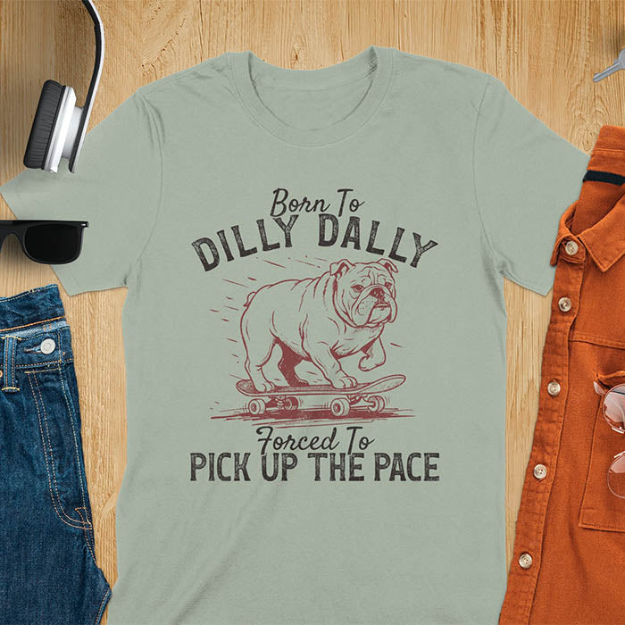 Grumpy bulldog on skateboard illustration with Born To Dilly Dally text and Forced To Pick Up The Pace slogan on sage green tee