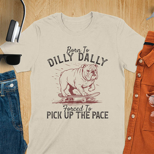 Grumpy bulldog on skateboard illustration with Born To Dilly Dally text and Forced To Pick Up The Pace slogan on sand color tee