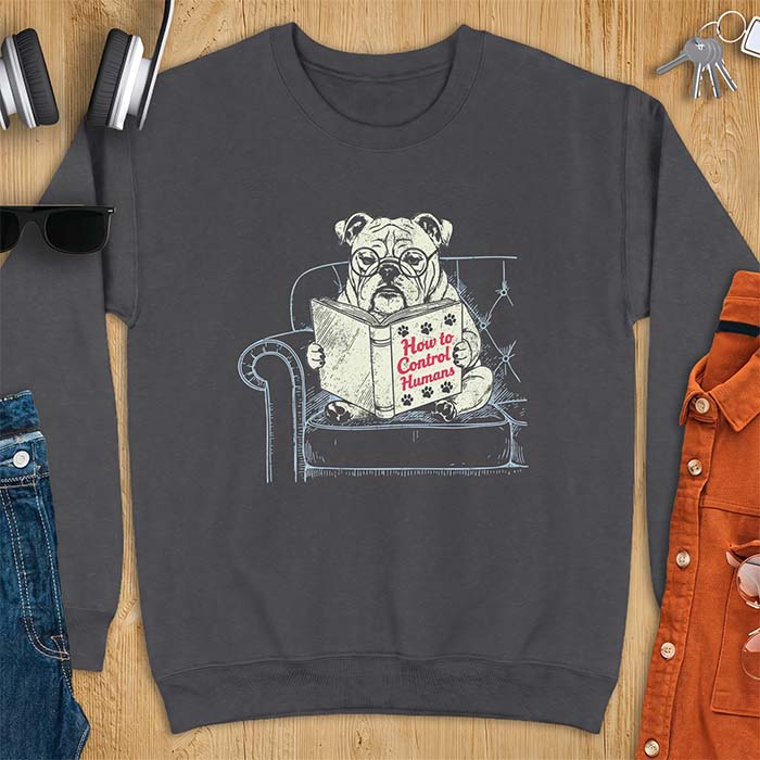 Dark Heather sweatshirt with a graphic of a bespectacled English Bulldog sitting in a tufted armchair reading a book titled How to Control Humans, decorated with paw prints