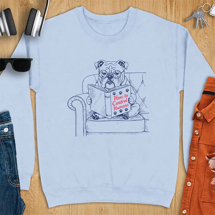 Light Blue sweatshirt with a graphic of a bespectacled English Bulldog sitting in a tufted armchair reading a book titled How to Control Humans, decorated with paw prints