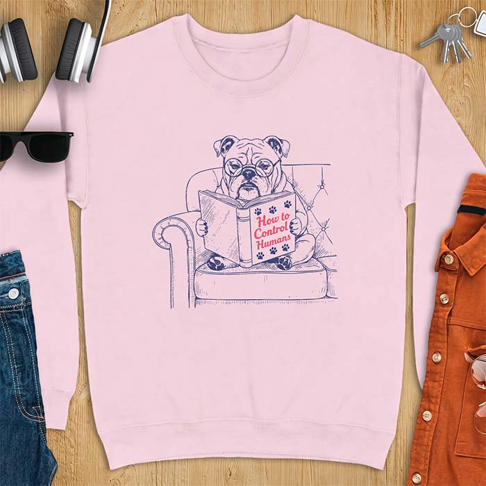 Light Pink sweatshirt with a graphic of a bespectacled English Bulldog sitting in a tufted armchair reading a book titled How to Control Humans, decorated with paw prints