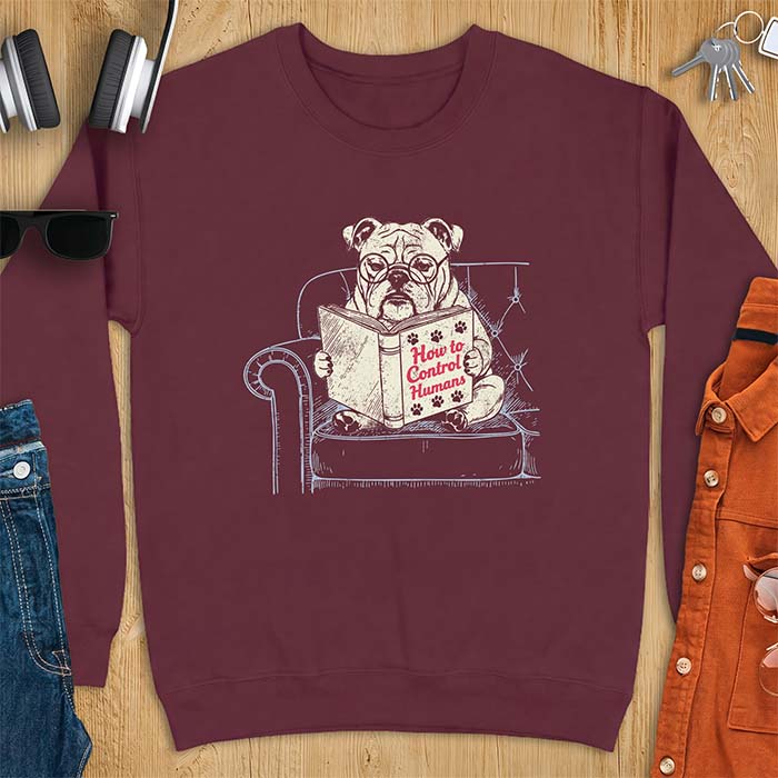 Maroon sweatshirt with a graphic of a bespectacled English Bulldog sitting in a tufted armchair reading a book titled How to Control Humans, decorated with paw prints