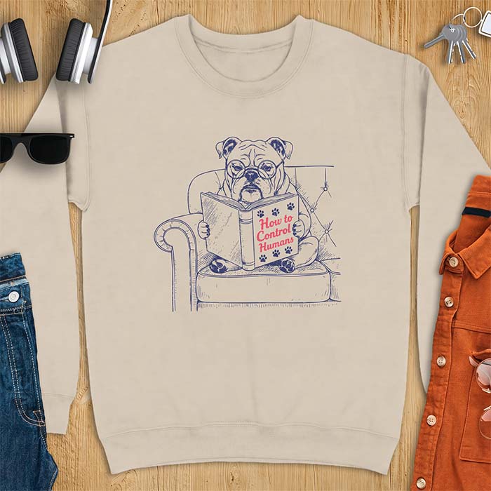 Sand sweatshirt with a graphic of a bespectacled English Bulldog sitting in a tufted armchair reading a book titled How to Control Humans, decorated with paw prints