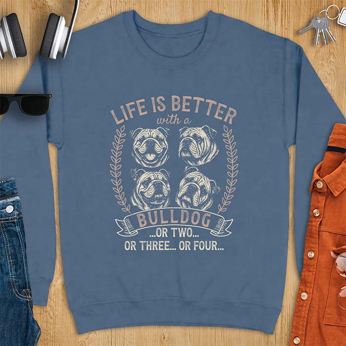 Indigo Blue sweatshirt with a graphic of four vintage-style bulldog head portraits framed by leafy wreaths and a ribbon banner reading Bulldog, featuring the text Life Is Better With A Bulldog, Or Two, Or Three, Or Four.