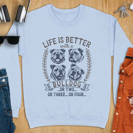 Light Blue sweatshirt with a graphic of four vintage-style bulldog head portraits framed by leafy wreaths and a ribbon banner reading Bulldog, featuring the text Life Is Better With A Bulldog, Or Two, Or Three, Or Four.