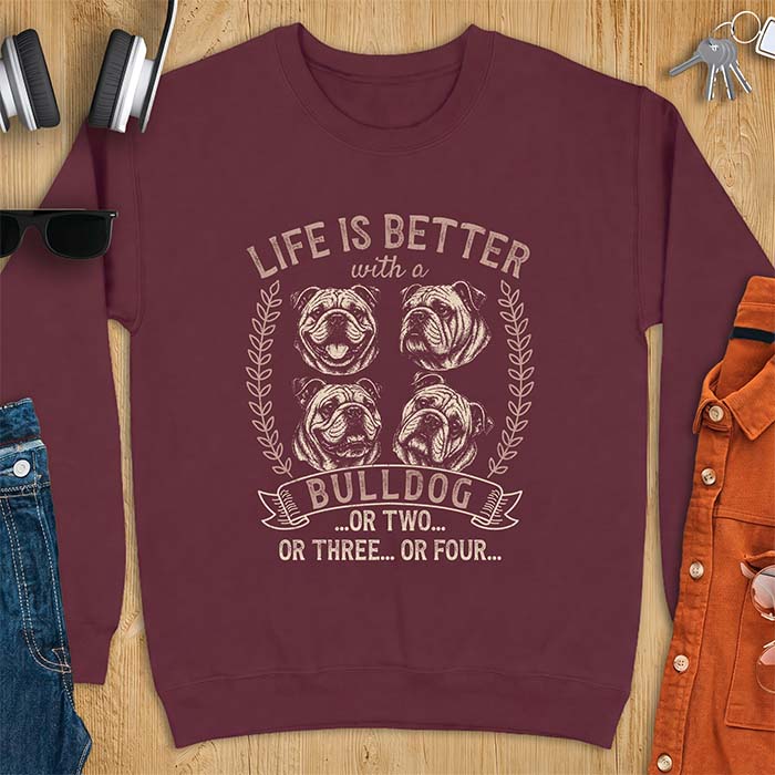 Maroon sweatshirt with a graphic of four vintage-style bulldog head portraits framed by leafy wreaths and a ribbon banner reading Bulldog, featuring the text Life Is Better With A Bulldog, Or Two, Or Three, Or Four.