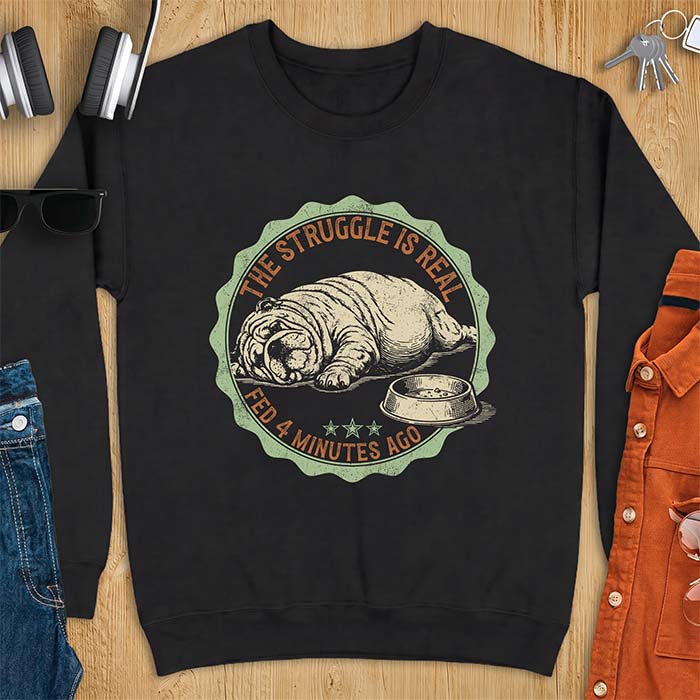 Black sweatshirt with a vintage badge graphic of an English Bulldog lying flat on the floor beside a nearly empty food bowl, with the text The Struggle Is Real above and Fed 4 Minutes Ago below