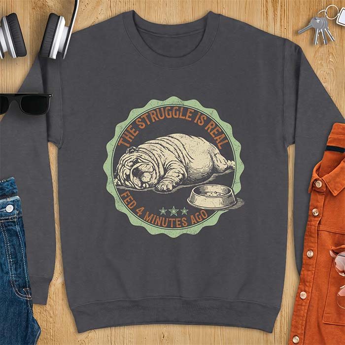 Dark heather sweatshirt with a vintage badge graphic of an English Bulldog lying flat on the floor beside a nearly empty food bowl, with the text The Struggle Is Real above and Fed 4 Minutes Ago below