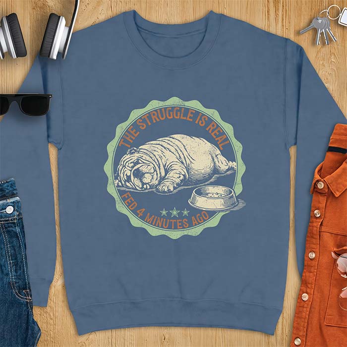 Indigo blue sweatshirt with a vintage badge graphic of an English Bulldog lying flat on the floor beside a nearly empty food bowl, with the text The Struggle Is Real above and Fed 4 Minutes Ago below