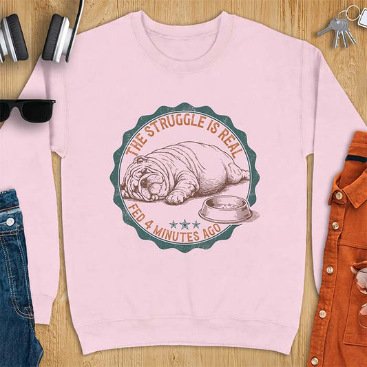 Light pink sweatshirt with a vintage badge graphic of an English Bulldog lying flat on the floor beside a nearly empty food bowl, with the text The Struggle Is Real above and Fed 4 Minutes Ago below