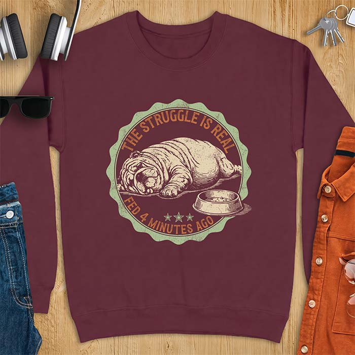 Maroon sweatshirt with a vintage badge graphic of an English Bulldog lying flat on the floor beside a nearly empty food bowl, with the text The Struggle Is Real above and Fed 4 Minutes Ago below