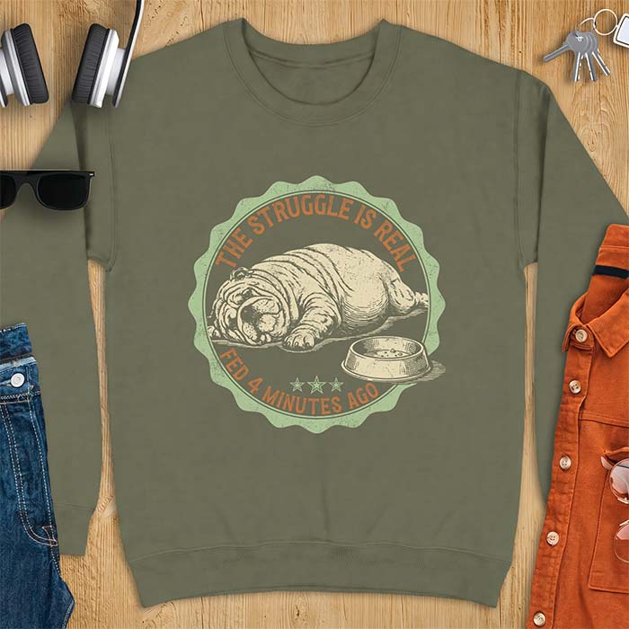 Military green sweatshirt with a vintage badge graphic of an English Bulldog lying flat on the floor beside a nearly empty food bowl, with the text The Struggle Is Real above and Fed 4 Minutes Ago below