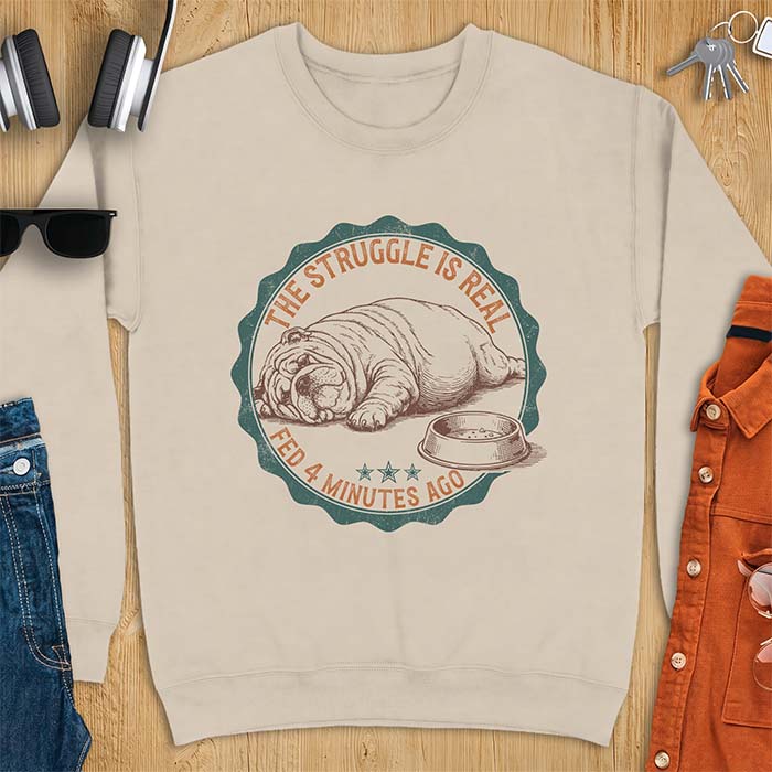 Sand sweatshirt with a vintage badge graphic of an English Bulldog lying flat on the floor beside a nearly empty food bowl, with the text The Struggle Is Real above and Fed 4 Minutes Ago below