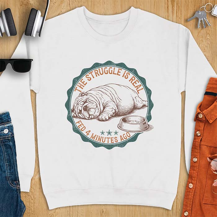 White sweatshirt with a vintage badge graphic of an English Bulldog lying flat on the floor beside a nearly empty food bowl, with the text The Struggle Is Real above and Fed 4 Minutes Ago below