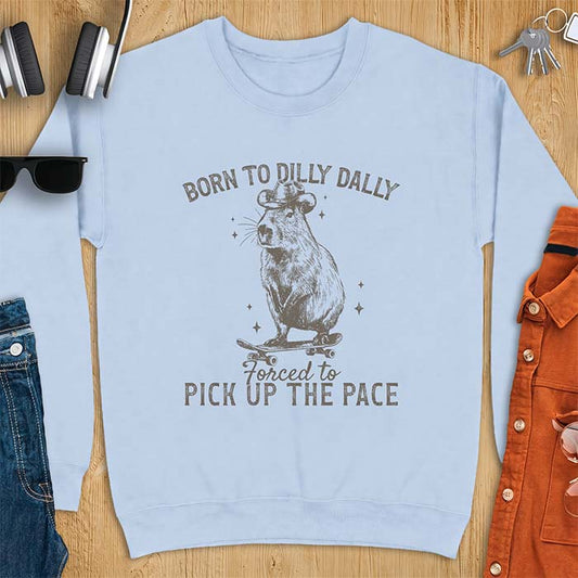 Light Blue sweatshirt with a graphic of a capybara wearing a cowboy hat riding a skateboard surrounded by sparkles, with the text Born To Dilly Dally Forced To Pick Up The Pace
