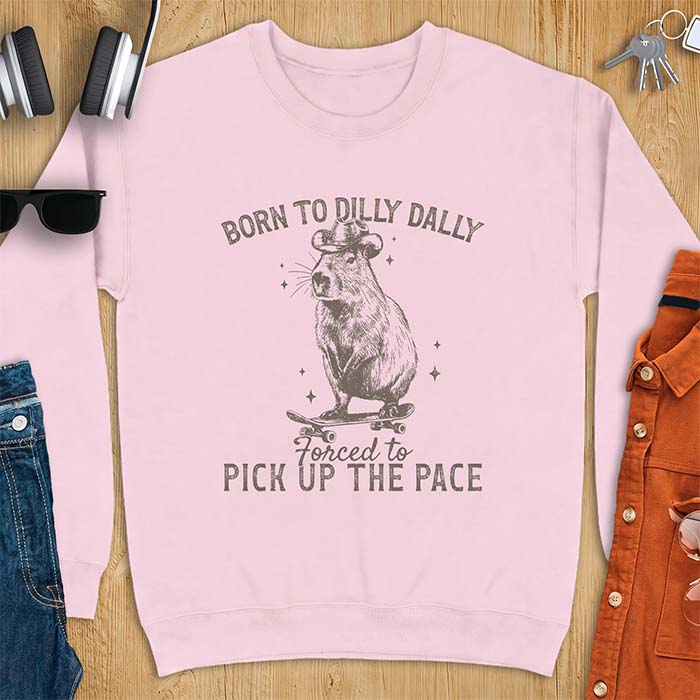 Light Pink sweatshirt with a graphic of a capybara wearing a cowboy hat riding a skateboard surrounded by sparkles, with the text Born To Dilly Dally Forced To Pick Up The Pace