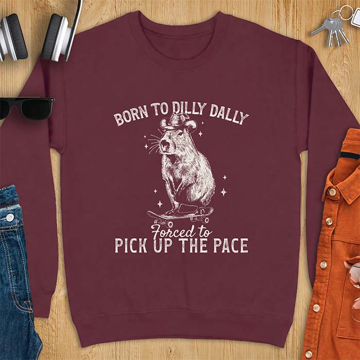 Maroon sweatshirt with a graphic of a capybara wearing a cowboy hat riding a skateboard surrounded by sparkles, with the text Born To Dilly Dally Forced To Pick Up The Pace