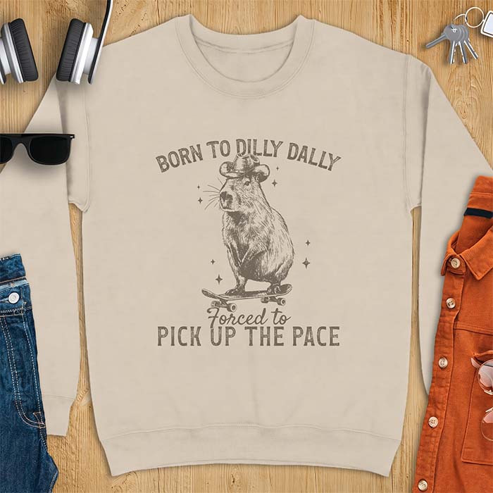 Sand sweatshirt with a graphic of a capybara wearing a cowboy hat riding a skateboard surrounded by sparkles, with the text Born To Dilly Dally Forced To Pick Up The Pace
