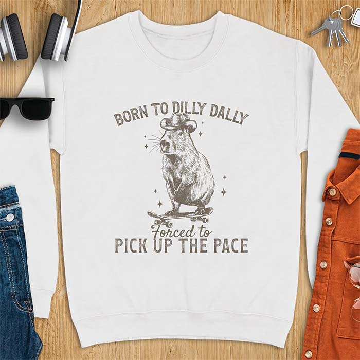 White sweatshirt with a graphic of a capybara wearing a cowboy hat riding a skateboard surrounded by sparkles, with the text Born To Dilly Dally Forced To Pick Up The Pace