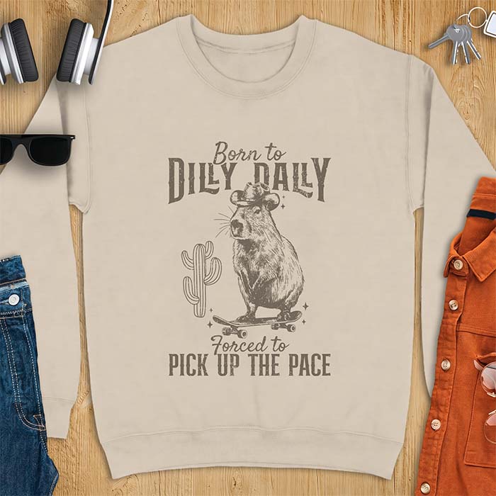 Sand sweatshirt with a graphic of a capybara wearing a cowboy hat balanced on a skateboard beside a cactus, plus the text Born to Dilly Dally and Forced to Pick Up The Pace.