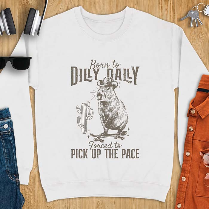 White sweatshirt with a graphic of a capybara wearing a cowboy hat balanced on a skateboard beside a cactus, plus the text Born to Dilly Dally and Forced to Pick Up The Pace.