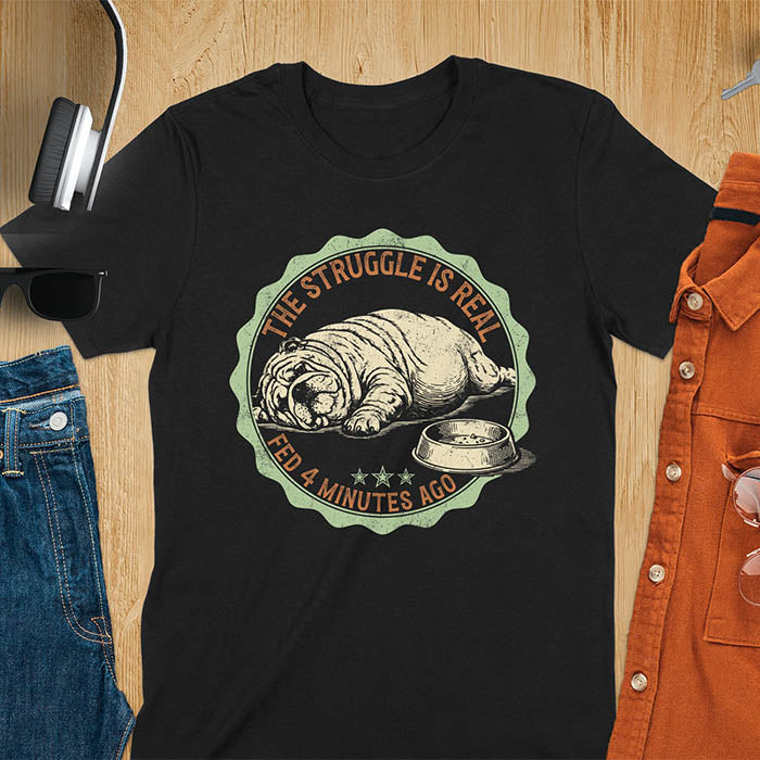 Black t-shirt with vintage badge graphic of a chubby English bulldog lying next to an empty food bowl, text reads "The Struggle Is Real Fed 4 Minutes Ago"