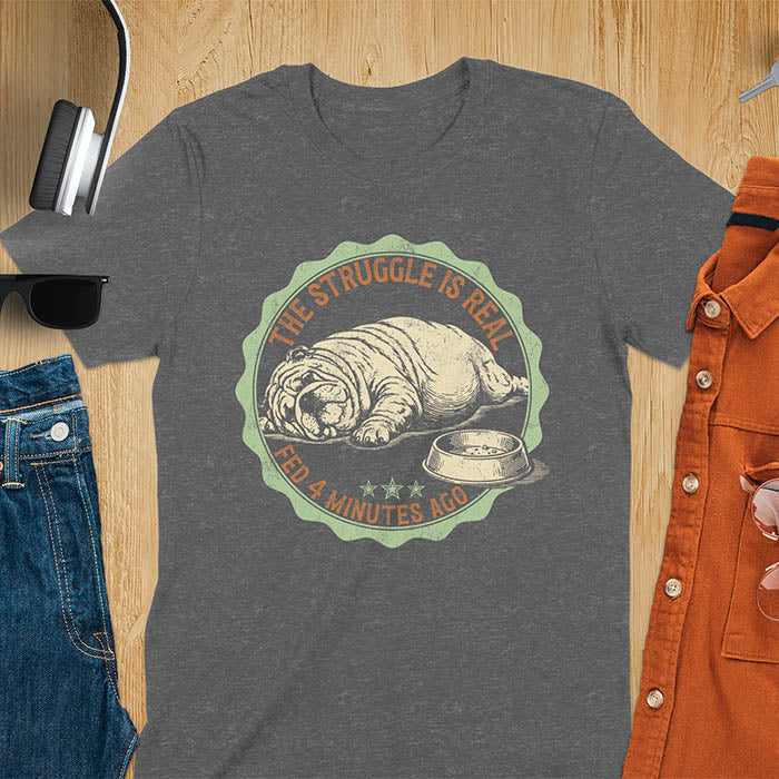 Dark heather t-shirt with vintage badge graphic of a chubby English bulldog lying next to an empty food bowl, text reads "The Struggle Is Real Fed 4 Minutes Ago"