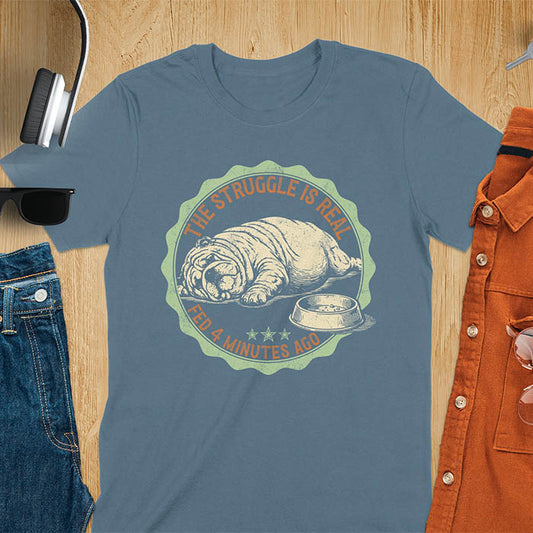 Indigo blue t-shirt with vintage badge graphic of a chubby English bulldog lying next to an empty food bowl, text reads "The Struggle Is Real Fed 4 Minutes Ago"