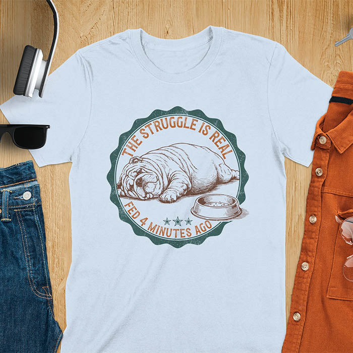 Light blue t-shirt with vintage badge graphic of a chubby English bulldog lying next to an empty food bowl, text reads "The Struggle Is Real Fed 4 Minutes Ago"