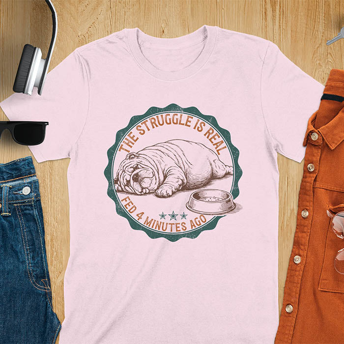 Light pink t-shirt with vintage badge graphic of a chubby English bulldog lying next to an empty food bowl, text reads "The Struggle Is Real Fed 4 Minutes Ago"