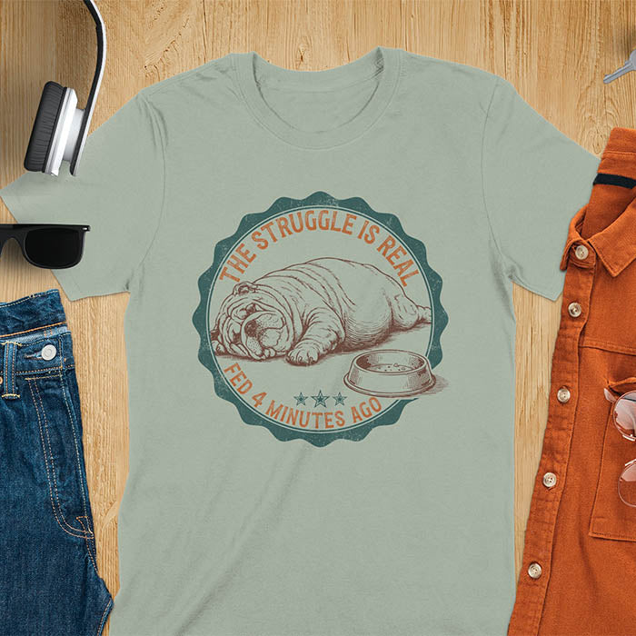 Sage green t-shirt with vintage badge graphic of a chubby English bulldog lying next to an empty food bowl, text reads "The Struggle Is Real Fed 4 Minutes Ago"