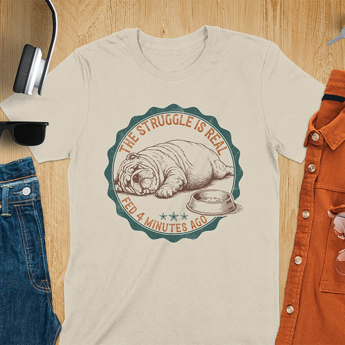 Sand color t-shirt with vintage badge graphic of a chubby English bulldog lying next to an empty food bowl, text reads "The Struggle Is Real Fed 4 Minutes Ago"