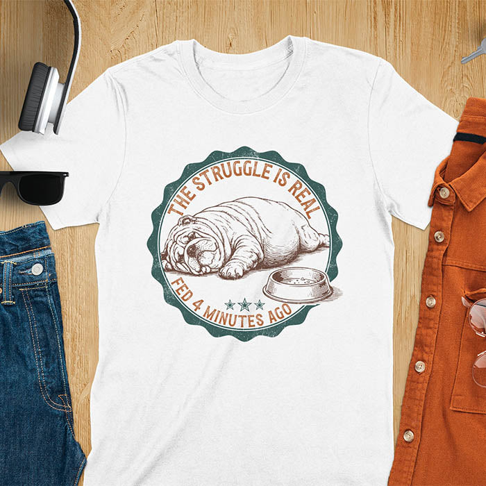 White t-shirt with vintage badge graphic of a chubby English bulldog lying next to an empty food bowl, text reads "The Struggle Is Real Fed 4 Minutes Ago"