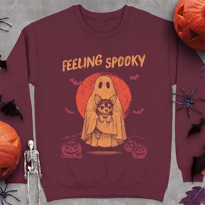 Maroon sweatshirt with a Halloween graphic of a ghost holding a wide-eyed Corgi puppy in front of a red moon with bats and jack-o-lanterns, with the text Feeling Spooky