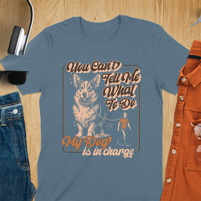 You Can't Tell Me What To Do My Dog Is In Charge text with man being walked on leash by corgi in vintage retro frame graphic on indigo blue t-shirt