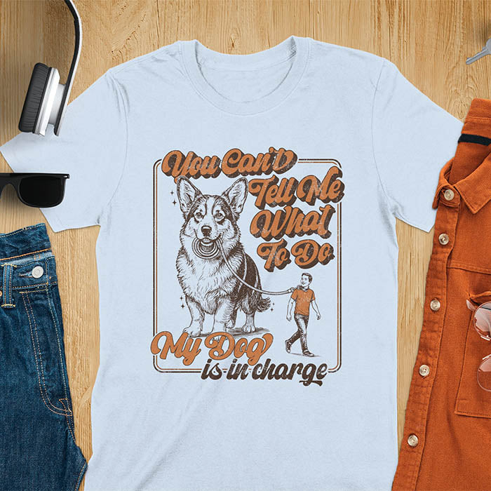 You Can't Tell Me What To Do My Dog Is In Charge text with man being walked on leash by corgi in vintage retro frame graphic on light blue t-shirt