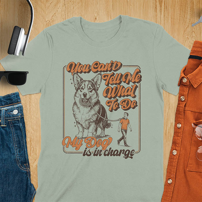 You Can't Tell Me What To Do My Dog Is In Charge text with man being walked on leash by corgi in vintage retro frame graphic on sage green t-shirt