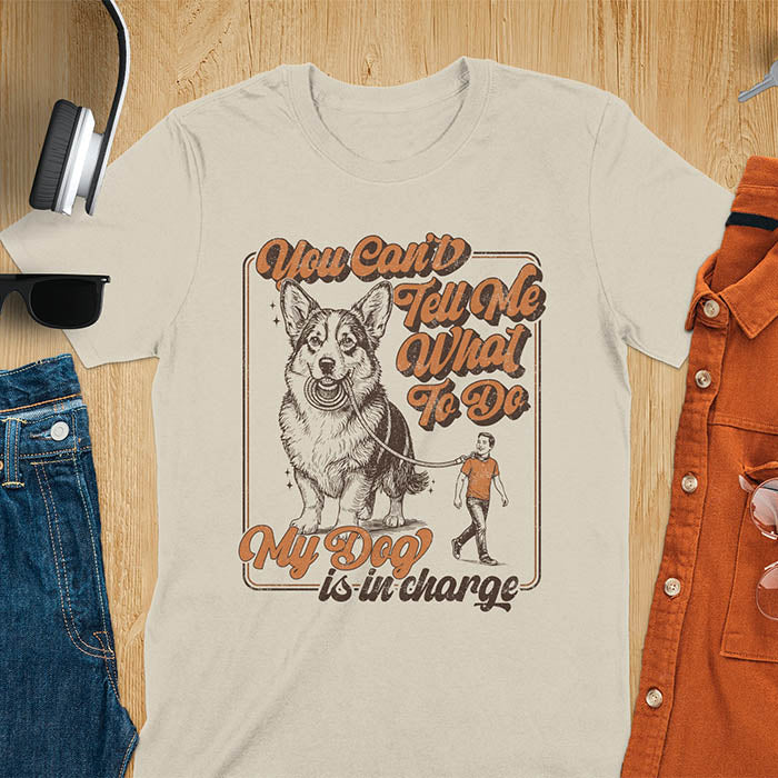 You Can't Tell Me What To Do My Dog Is In Charge text with man being walked on leash by corgi in vintage retro frame graphic on sand color t-shirt