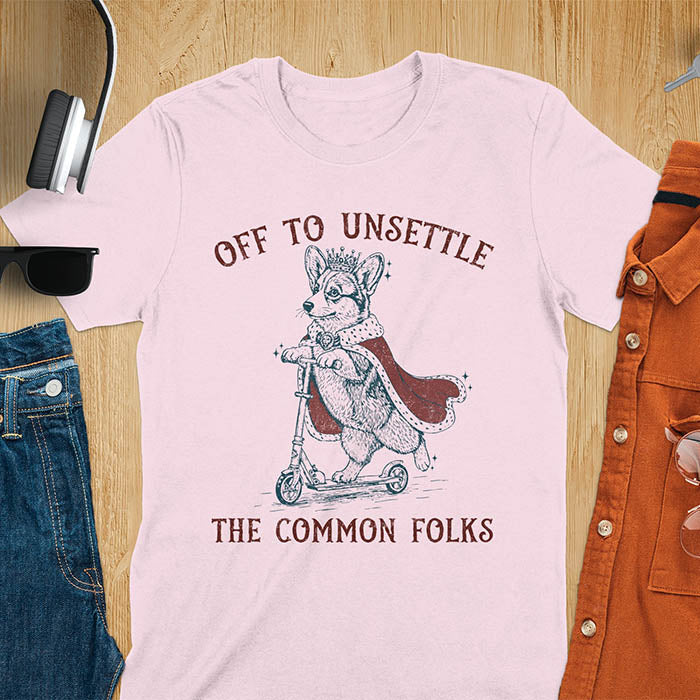 Off To Unsettle The Common Folks text with king corgi wearing crown and red cape on skateboard scooter vintage meme graphic on light pink t-shirt