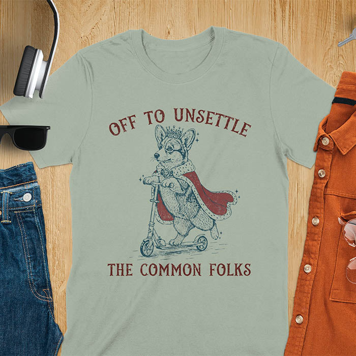 Off To Unsettle The Common Folks text with king corgi wearing crown and red cape on skateboard scooter vintage meme graphic on sage green t-shirt
