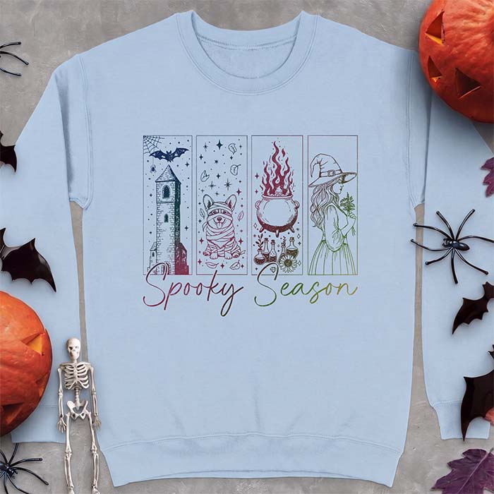 Light blue sweatshirt with a four-panel Halloween graphic of a haunted tower, a bandage-wrapped Corgi mummy, a bubbling cauldron with potion bottles and a witch holding flowers, with the text Spooky Season