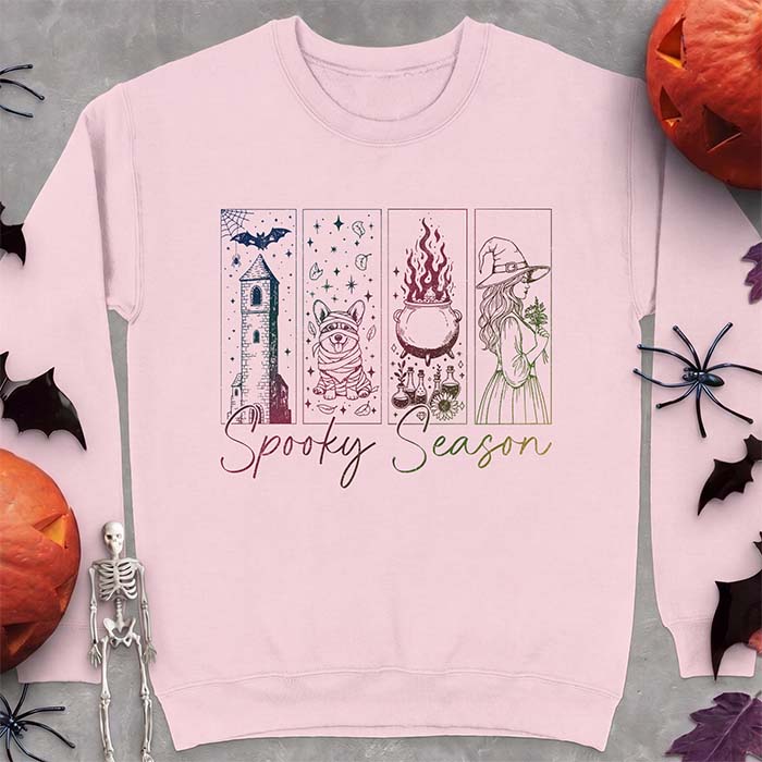 A light pink sweatshirt with a four-panel Halloween graphic of a haunted tower, a bandage-wrapped Corgi mummy, a bubbling cauldron with potion bottles and a witch holding flowers, with the text Spooky Season