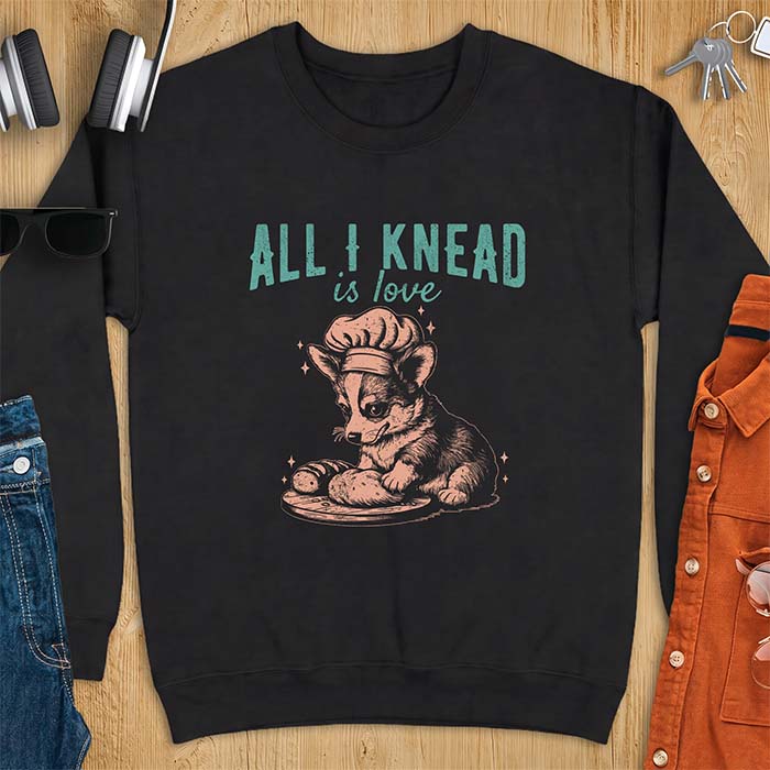 Black sweatshirt with a graphic of a corgi puppy wearing a chef's hat kneading a loaf of bread at a cutting board with sparkles around it, featuring the text All I Knead Is Love.